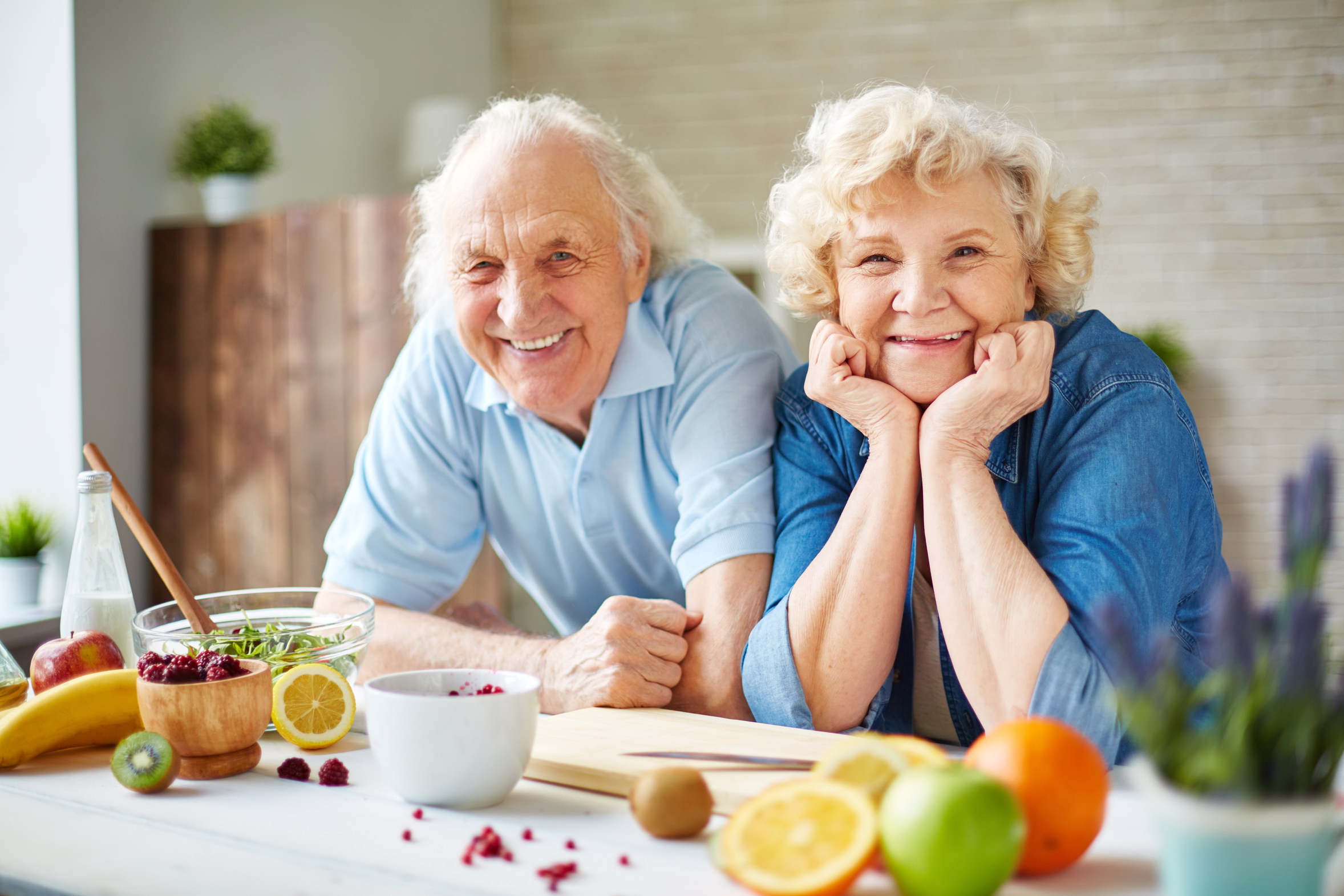 Zwei Senioren lehnen auf einem Küchentisch auf dem Früchte liegen.