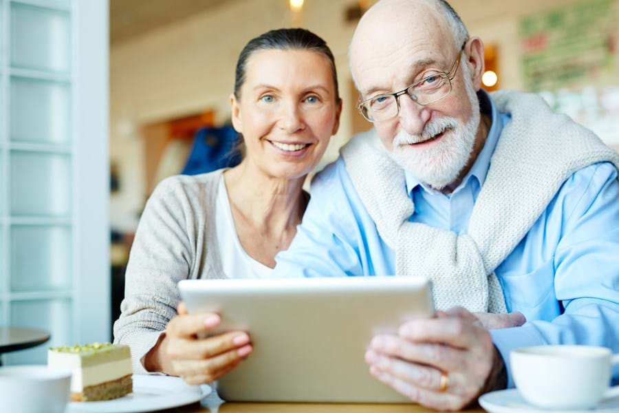 Zwei Senioren sitzen am Tisch und halten gemeinsam ein silbernes Tablet-Geräte in den Händen