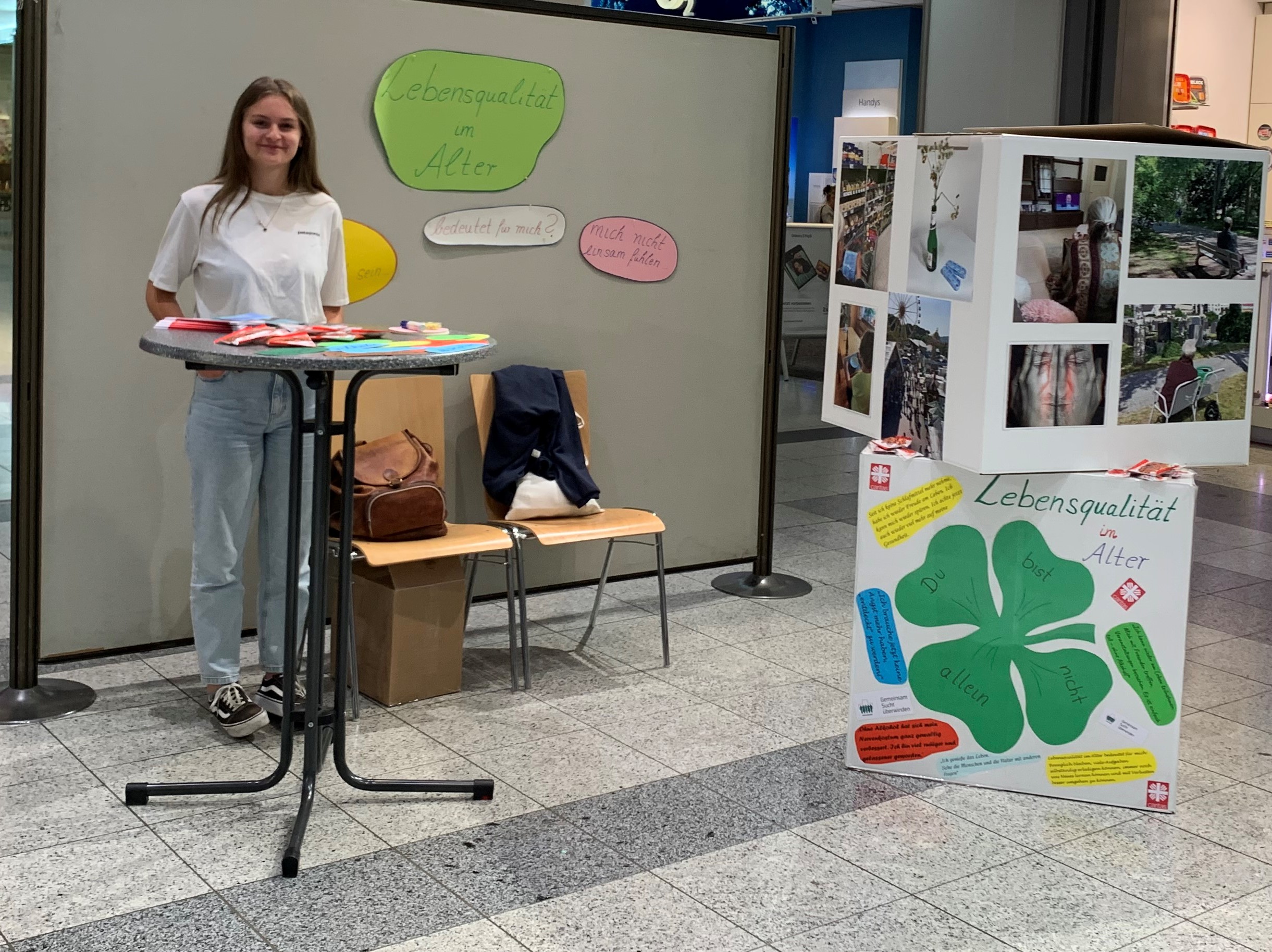 Mitarbeiterin steht vor dem Stand der Caritas-Suchtambulanz Regensburg bestehend aus einer Pinnwand und zwei selbstgebastelten Würfeln mit Fotos und Fragen zur Lebensqualität.